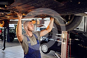 Handsome auto mechanic checking running gear of automobile on service station. Cheerful male worker fixing problem with car.