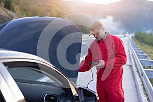 Handsome auto mechanic or road assistance worker in uniform repairing engine of the broken car on the road