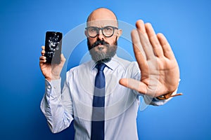 Handsome bald business man with beard holding broken smartphone showing cracked screen with open hand doing stop sign with serious