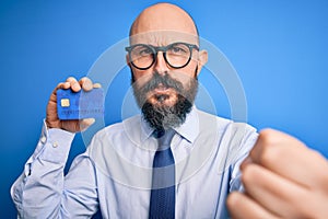 Handsome bald business man with beard holding credit card over isolated blue background annoyed and frustrated shouting with