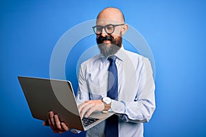 Handsome bald business man with beard working using laptop over blue background with a happy face standing and smiling with a