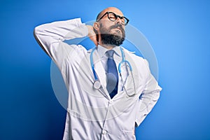 Handsome bald doctor man with beard wearing glasses and stethoscope over blue background Suffering of neck ache injury, touching