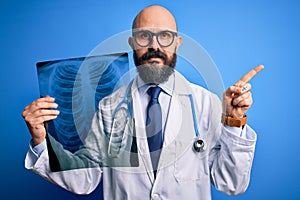 Handsome bald doctor man with beard wearing stethoscope holding chest xray very happy pointing with hand and finger to the side