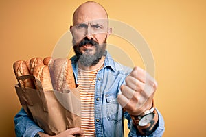 Handsome bald man with beard holding paper bag with bread over yellow background annoyed and frustrated shouting with anger, crazy