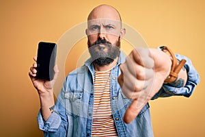 Handsome bald man with beard holding smartphone showing screen over yellow background with angry face, negative sign showing