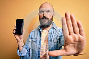 Handsome bald man with beard holding smartphone showing screen over yellow background with open hand doing stop sign with serious