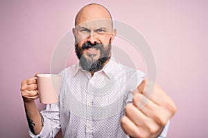 Handsome bald man with beard and tattoo drinking cup of coffee over pink background annoyed and frustrated shouting with anger,