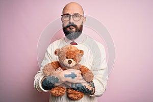 Handsome bald man with beard and tattoo holding teddy bear over isolated pink background with a confident expression on smart face