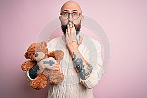 Handsome bald man with beard and tattoo holding teddy bear over isolated pink background cover mouth with hand shocked with shame