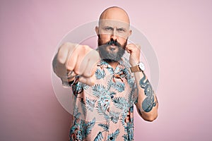 Handsome bald man with beard and tattoo wearing casual floral shirt over pink background Punching fist to fight, aggressive and