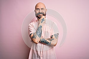 Handsome bald man with beard and tattoo wearing casual shirt over isolated pink background looking confident at the camera smiling
