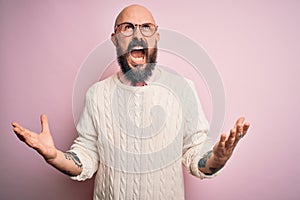 Handsome bald man with beard and tattoo wearing glasses and sweater over pink background crazy and mad shouting and yelling with