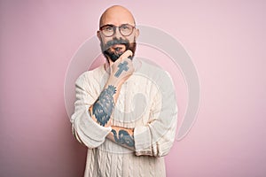 Handsome bald man with beard and tattoo wearing glasses and sweater over pink background looking confident at the camera smiling