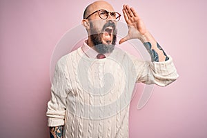 Handsome bald man with beard and tattoo wearing glasses and sweater over pink background shouting and screaming loud to side with