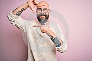 Handsome bald man with beard and tattoo wearing glasses and sweater over pink background smiling making frame with hands and