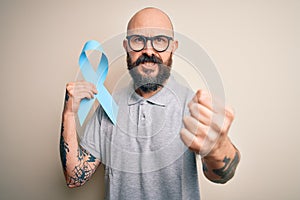 Handsome bald man with beard and tattoos holding blue cancer ribbon over isolated background annoyed and frustrated shouting with