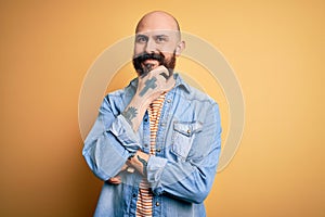 Handsome bald man with beard wearing casual denim jacket and striped t-shirt looking confident at the camera smiling with crossed