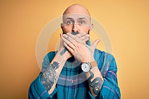 Handsome bald man with beard wearing casual shirt standing over  yellow background shocked covering mouth with hands for