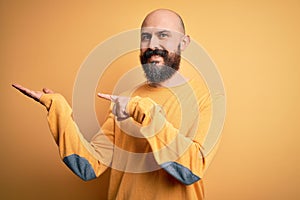 Handsome bald man with beard wearing casual sweater standing over yellow background amazed and smiling to the camera while