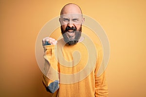 Handsome bald man with beard wearing casual sweater standing over yellow background angry and mad raising fist frustrated and
