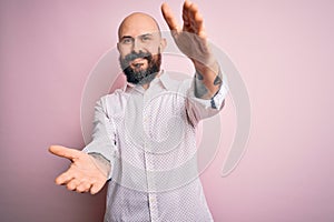 Handsome bald man with beard wearing elegant shirt over isolated pink background looking at the camera smiling with open arms for