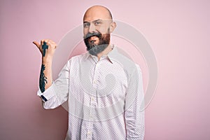 Handsome bald man with beard wearing elegant shirt over isolated pink background smiling with happy face looking and pointing to