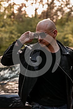 Handsome bald unshaved man in fashionable clothes stands and straightens sunglasses near black car at sunset on nature. Cool guy