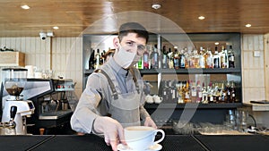 Handsome barista in protective mask giving a coffee to the client at the bar of the modern cafe