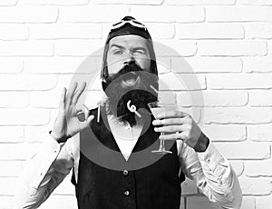 Handsome bearded aviator man with long beard and mustache on funny face holding glass of alcoholic cocktail in vintage