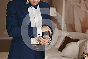 Handsome bearded brutal man in a stylish blue suit and in bowtie puts on stylish cufflinks and posing at interior