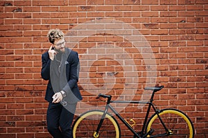 Handsome bearded businessman in classic suit is talk a smart phone and smiling while riding bicycle standing against brick wall