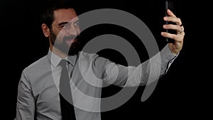 Handsome bearded businessman on isolated black background greets in a video call on cellphone.