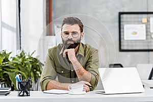 handsome bearded businessman resting chin on hand and looking at camera