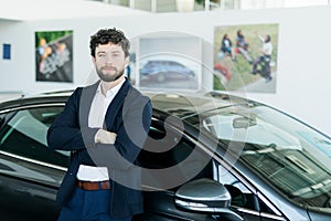 Handsome bearded buyer in casual wear in dealership, guy looks on camera while standing near car with crosed arms