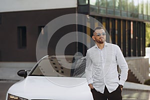 Handsome bearded buyer in casual wear in dealership, guy looks on camera while standing near car with crosed arms.