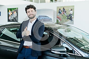 Handsome bearded buyer in casual wear in dealership, guy looks on camera while standing near car with thumbs up