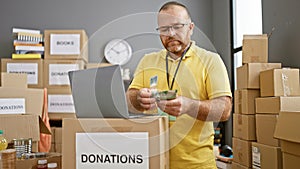 Handsome bearded caucasian man volunteering at a charity center, seriously counting australian dollars on laptop amidst boxes of