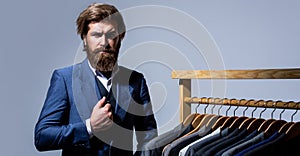 Handsome bearded fashion man in classical costume suit. Male suits hanging in a row. Men clothing, boutiques. Tailor