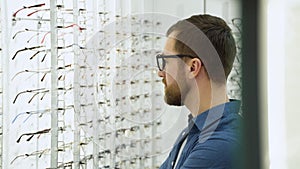 Handsome bearded guy picking new glasses at optical shop, looking at mirror