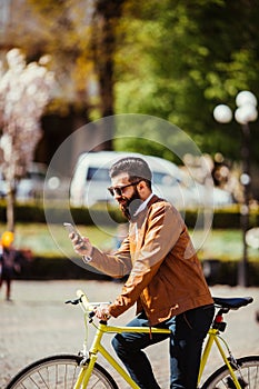 Handsome bearded hipster man is using a smart phone and smiling while riding bicycle in city