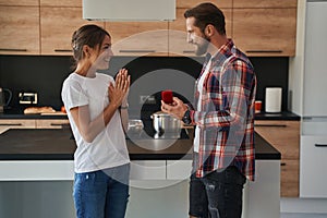 Handsome bearded male with engagement ring in a box and happy cheerful lady in their house