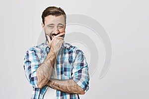 Handsome bearded man covering mouth with hand trying not to laught out loud over something, standing against gray wall photo