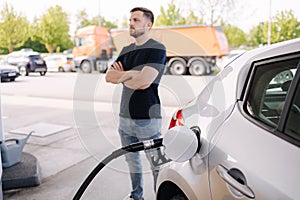 Handsome bearded man cross the arms during refueling car. Male looking on the scoreboard at gas station