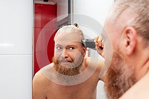 Handsome bearded man cutting his own hair with hair clipper in bathroom in front of mirror at home. Closeup