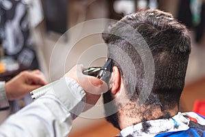 Handsome bearded man, getting haircut by barber, with electric trimmer at barbershop.