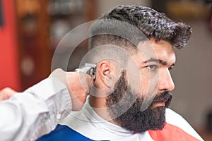 Handsome bearded man, getting haircut by barber, with electric trimmer at barbershop.