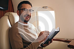 handsome bearded man in glasses looking at camera while holding notebook in private jet.