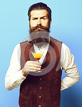Handsome bearded man with long beard and mustache has stylish hair on serious face holding glass of alcoholic cocktail