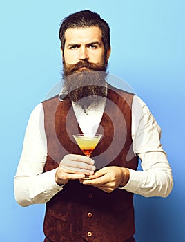 Handsome bearded man with long beard and mustache has stylish hair on serious face holding glass of alcoholic cocktail