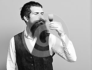 Handsome bearded man with long beard and mustache on serious face tasting glass of alcoholic shot in vintage suede